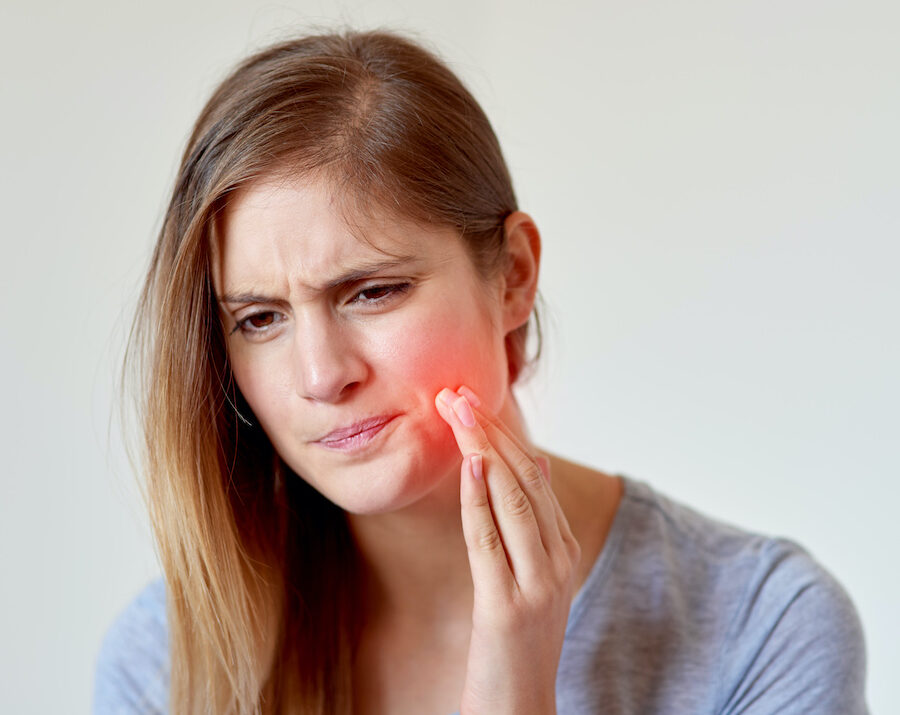 Blonde woman cringes in pain and touches a red hotspot on her cheek due to underlying tooth pain that needs root canal treatment