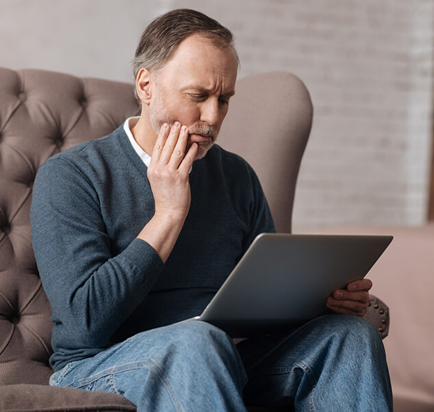 man looking at his laptop while holding his jaw in pain