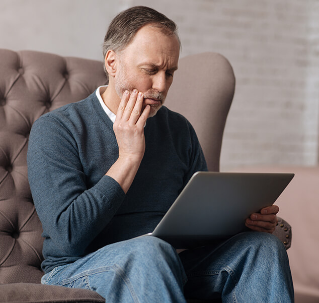 man holding his jaw in pain while looking at a laptop